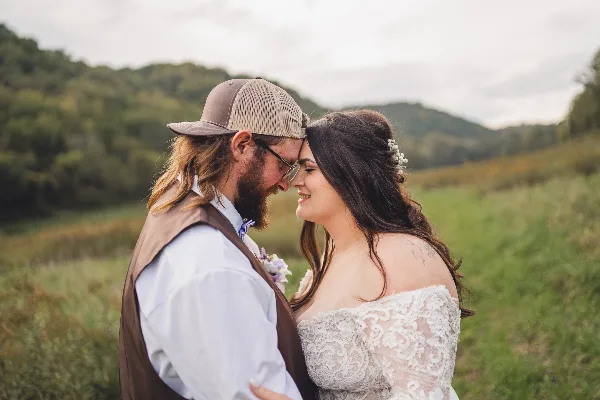 A bride and groom standing close together in a grassy field with a forested hill in the background.