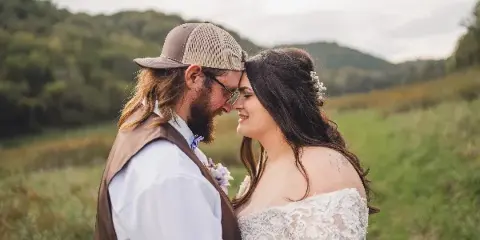 A bride and groom standing close together in a grassy field with a forested hill in the background.