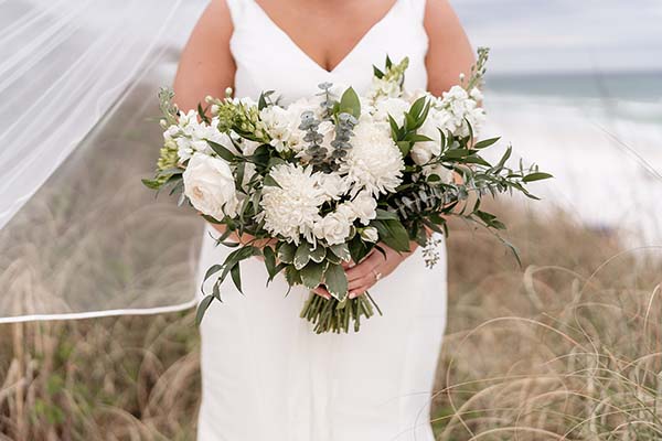 A bride in a white dress holding a large bouquet of white flowers and greenery on a beach.