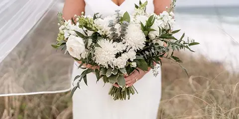 A bride in a white dress holding a large bouquet of white flowers and greenery on a beach.
