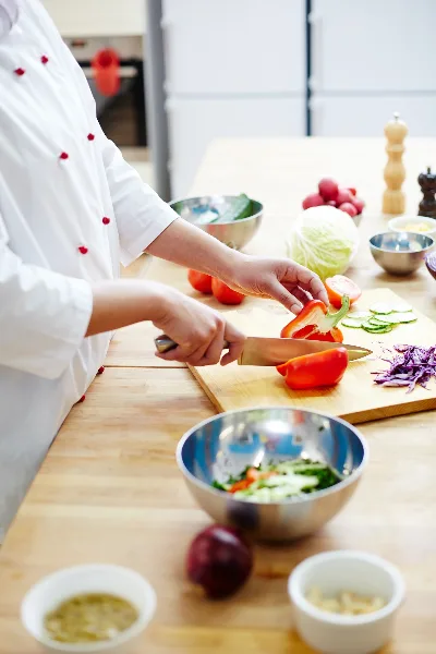 A chef in a white uniform slicing red peppers on a wooden cutting board in a kitchen.