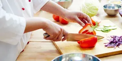 A chef in a white uniform slicing red peppers on a wooden cutting board in a kitchen.