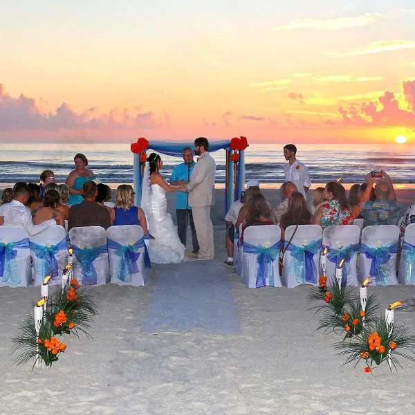 A bride and groom exchanging vows on a beach at sunset with seated guests and a decorated arch.