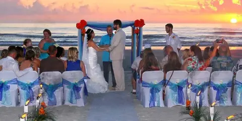A bride and groom exchanging vows on a beach at sunset with seated guests and a decorated arch.