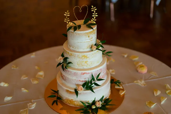 A four-tier semi-naked wedding cake decorated with fresh greenery, small pink roses, and a gold heart topper, sitting on a gold cake stand.