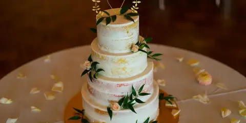 A four-tier semi-naked wedding cake decorated with fresh greenery, small pink roses, and a gold heart topper, sitting on a gold cake stand.