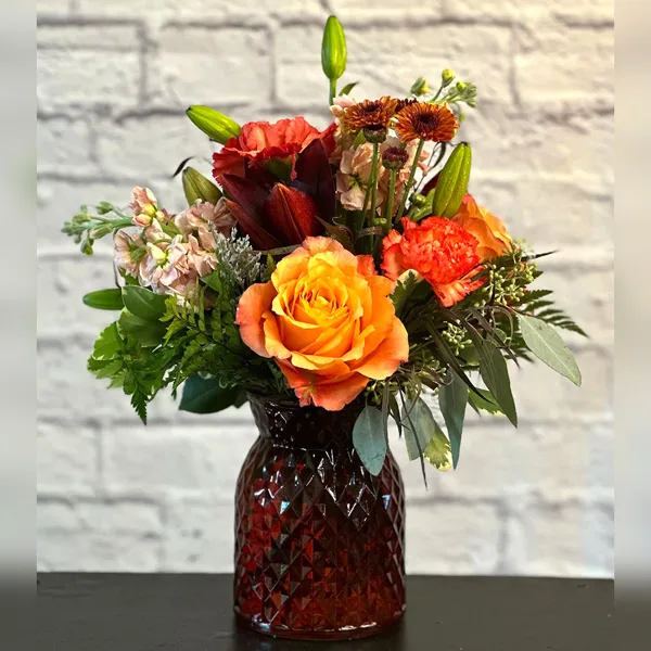 A floral arrangement with orange roses, red lilies, and greenery in a textured amber glass vase.