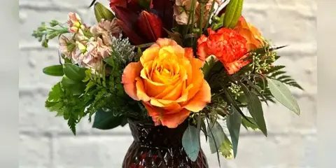 A floral arrangement with orange roses, red lilies, and greenery in a textured amber glass vase.
