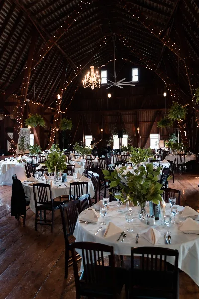 Interior of a rustic barn wedding venue featuring round tables with white linens, dark wooden chairs, and string lights hanging from the high ceiling.