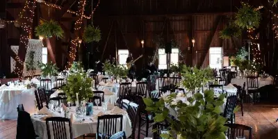 Interior of a rustic barn wedding venue featuring round tables with white linens, dark wooden chairs, and string lights hanging from the high ceiling.