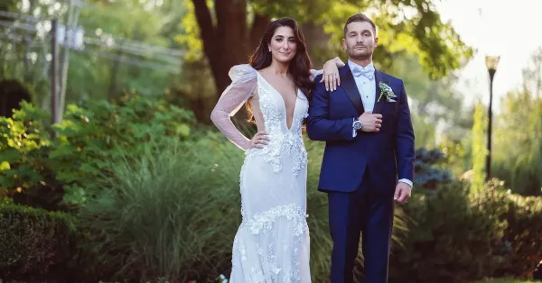 A bride in a white lace gown and a groom in a navy suit standing together outdoors in a garden setting.