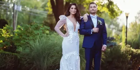 A bride in a white lace gown and a groom in a navy suit standing together outdoors in a garden setting.