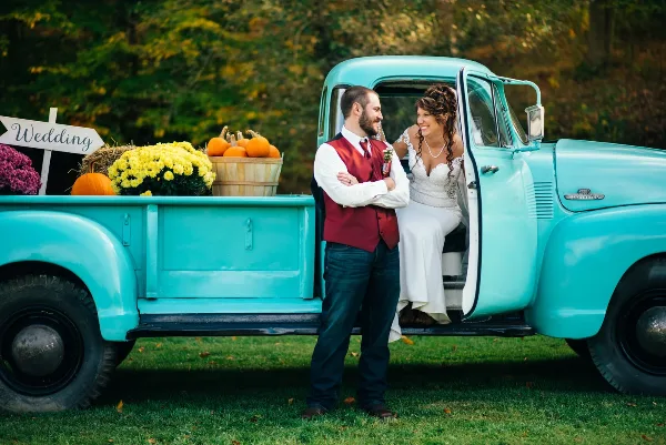 A bride and groom posing next to a vintage turquoise pickup truck decorated with pumpkins and flowers.