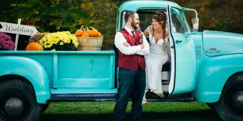 A bride and groom posing next to a vintage turquoise pickup truck decorated with pumpkins and flowers.