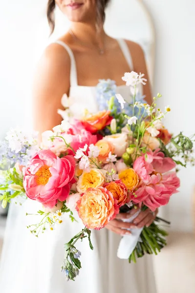 A close-up of a colorful bridal bouquet with pink peonies and orange ranunculus held by a bride in a white dress.