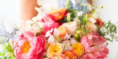 A close-up of a colorful bridal bouquet with pink peonies and orange ranunculus held by a bride in a white dress.