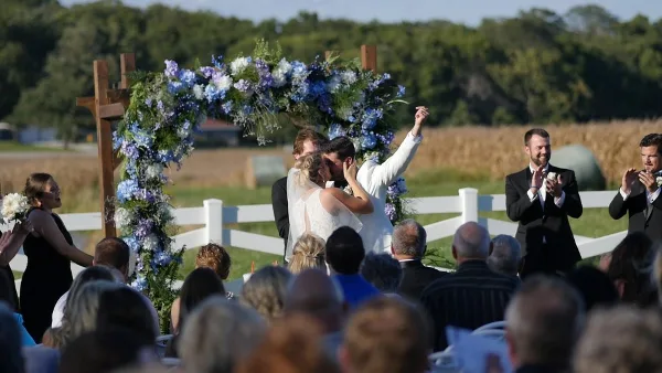 Outdoor wedding ceremony with a floral arch, bride and groom, and guests seated in a field.