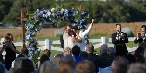 Outdoor wedding ceremony with a floral arch, bride and groom, and guests seated in a field.