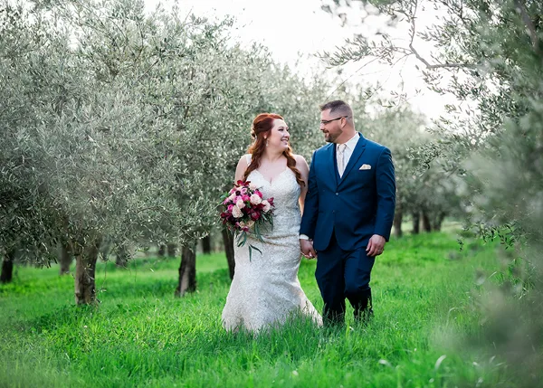 A bride in a white lace gown holding a bouquet and a groom in a blue suit walking through a grassy olive grove.