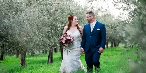 A bride in a white lace gown holding a bouquet and a groom in a blue suit walking through a grassy olive grove.