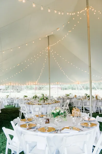 A wedding reception venue featuring a white tent with string lights and round tables set with white linens, gold chargers, and floral centerpieces.