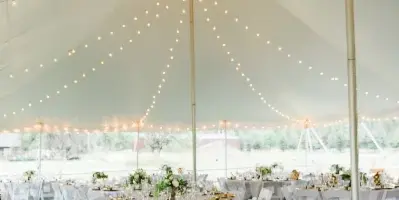 A wedding reception venue featuring a white tent with string lights and round tables set with white linens, gold chargers, and floral centerpieces.