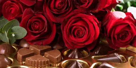 A close-up photograph of a bouquet of red roses arranged with chocolate candies.