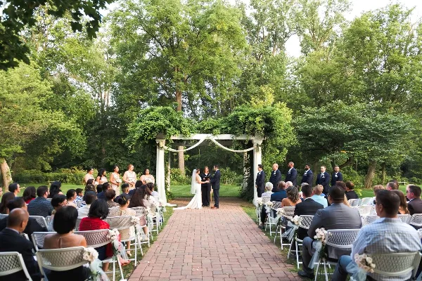 A wide shot of an outdoor wedding ceremony with a bride and groom standing at an altar under a green arbor, surrounded by seated guests and a wedding party.