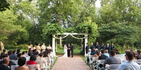 A wide shot of an outdoor wedding ceremony with a bride and groom standing at an altar under a green arbor, surrounded by seated guests and a wedding party.
