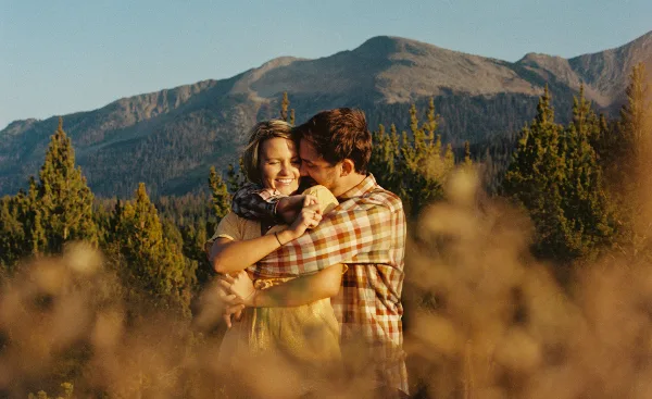 A cinematic film still of a couple embracing in a mountainous landscape with pine trees and a mountain peak in the background.