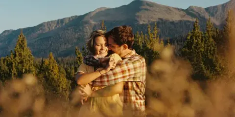 A cinematic film still of a couple embracing in a mountainous landscape with pine trees and a mountain peak in the background.