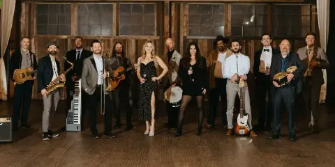 A group of musicians standing in a line holding various instruments including guitars, saxophones, and drums.
