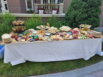 A long table covered with a white cloth, filled with numerous catering dishes, platters, and food items set up outdoors.