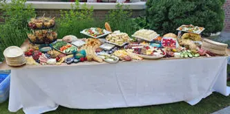 A long table covered with a white cloth, filled with numerous catering dishes, platters, and food items set up outdoors.