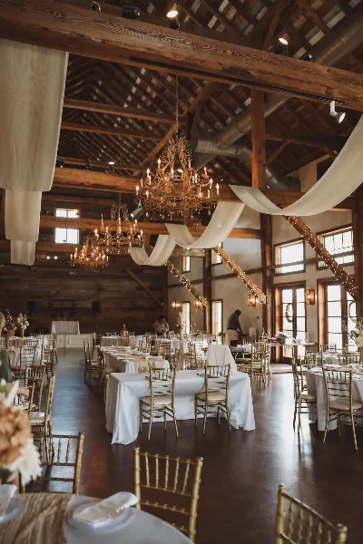 Interior of a rustic wooden venue set up for a wedding reception with white table linens, gold chiavari chairs, and hanging chandeliers.