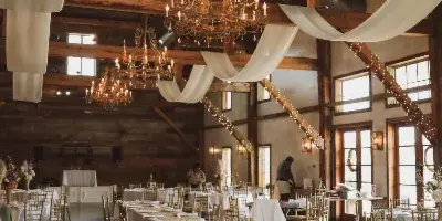 Interior of a rustic wooden venue set up for a wedding reception with white table linens, gold chiavari chairs, and hanging chandeliers.