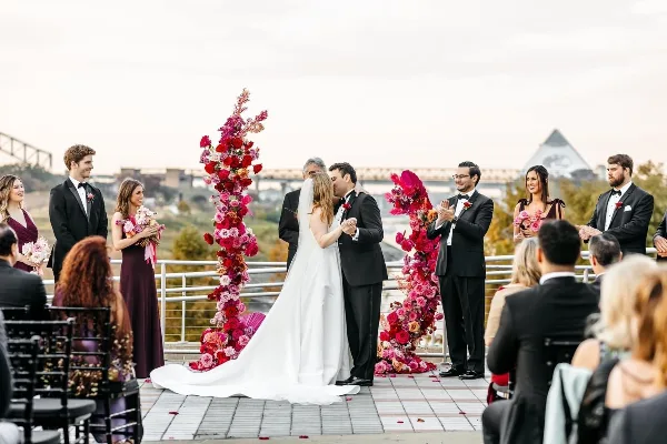 A bride and groom sharing a kiss during their wedding ceremony on an outdoor terrace with floral arches and wedding party members.