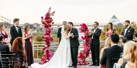 A bride and groom sharing a kiss during their wedding ceremony on an outdoor terrace with floral arches and wedding party members.