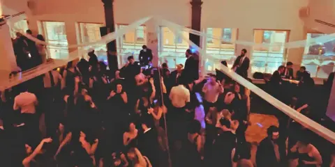 A crowded dance floor at a wedding event with a DJ at the booth and guests dancing.