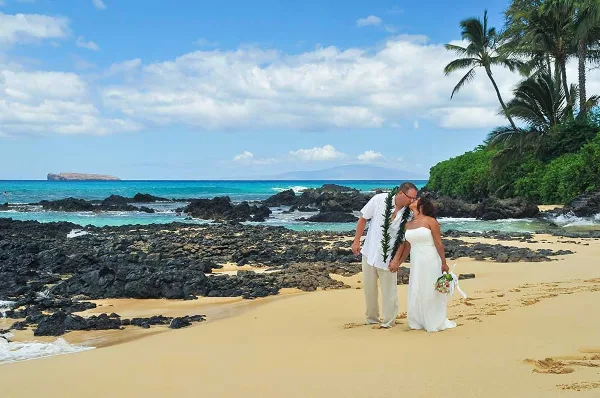 A bride and groom wearing leis standing on a sandy beach with palm trees and the ocean in the background.