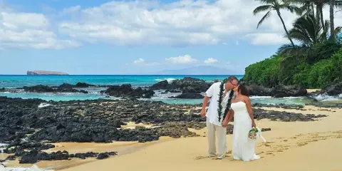 A bride and groom wearing leis standing on a sandy beach with palm trees and the ocean in the background.
