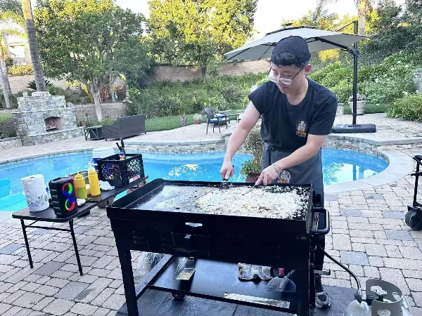 A chef in a black shirt and apron cooking rice on a portable hibachi grill next to a swimming pool.