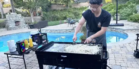 A chef in a black shirt and apron cooking rice on a portable hibachi grill next to a swimming pool.