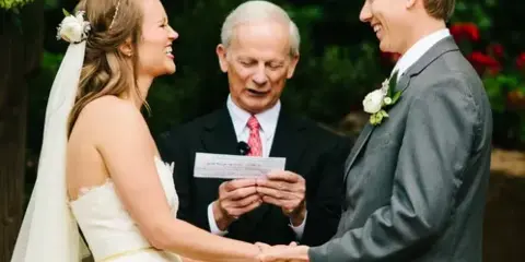 A bride and groom holding hands during an outdoor wedding ceremony with an officiant.