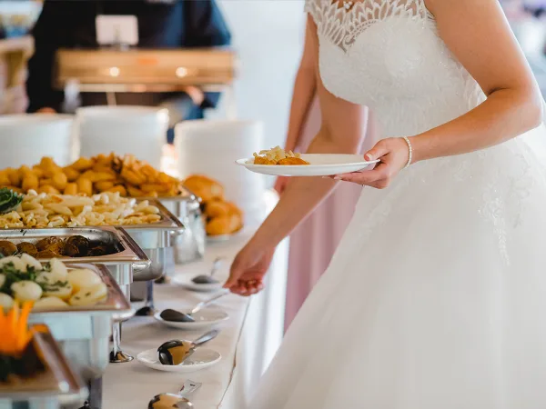 A bride in a white lace dress holding a plate of food while standing in front of a catering buffet with chafing dishes.
