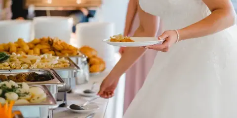 A bride in a white lace dress holding a plate of food while standing in front of a catering buffet with chafing dishes.