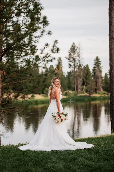 A bride in a white wedding dress holding a bouquet stands on a grassy riverbank with pine trees in the background.