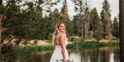 A bride in a white wedding dress holding a bouquet stands on a grassy riverbank with pine trees in the background.