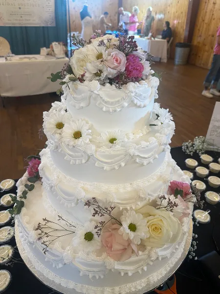 A four-tier white wedding cake decorated with white and pink flowers and buttercream piping, displayed on a table at an event venue.