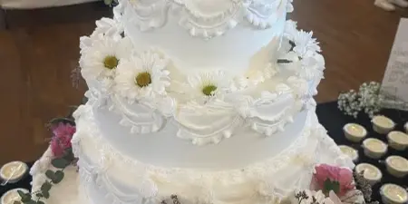 A four-tier white wedding cake decorated with white and pink flowers and buttercream piping, displayed on a table at an event venue.
